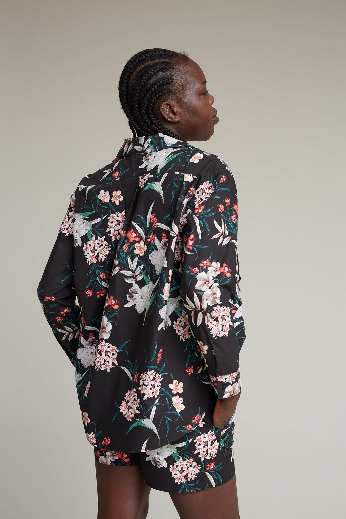 A person with braided hair is facing to the side, wearing a black long-sleeve loungewear set adorned with a vibrant tropical flower print. The background is a plain light grey.