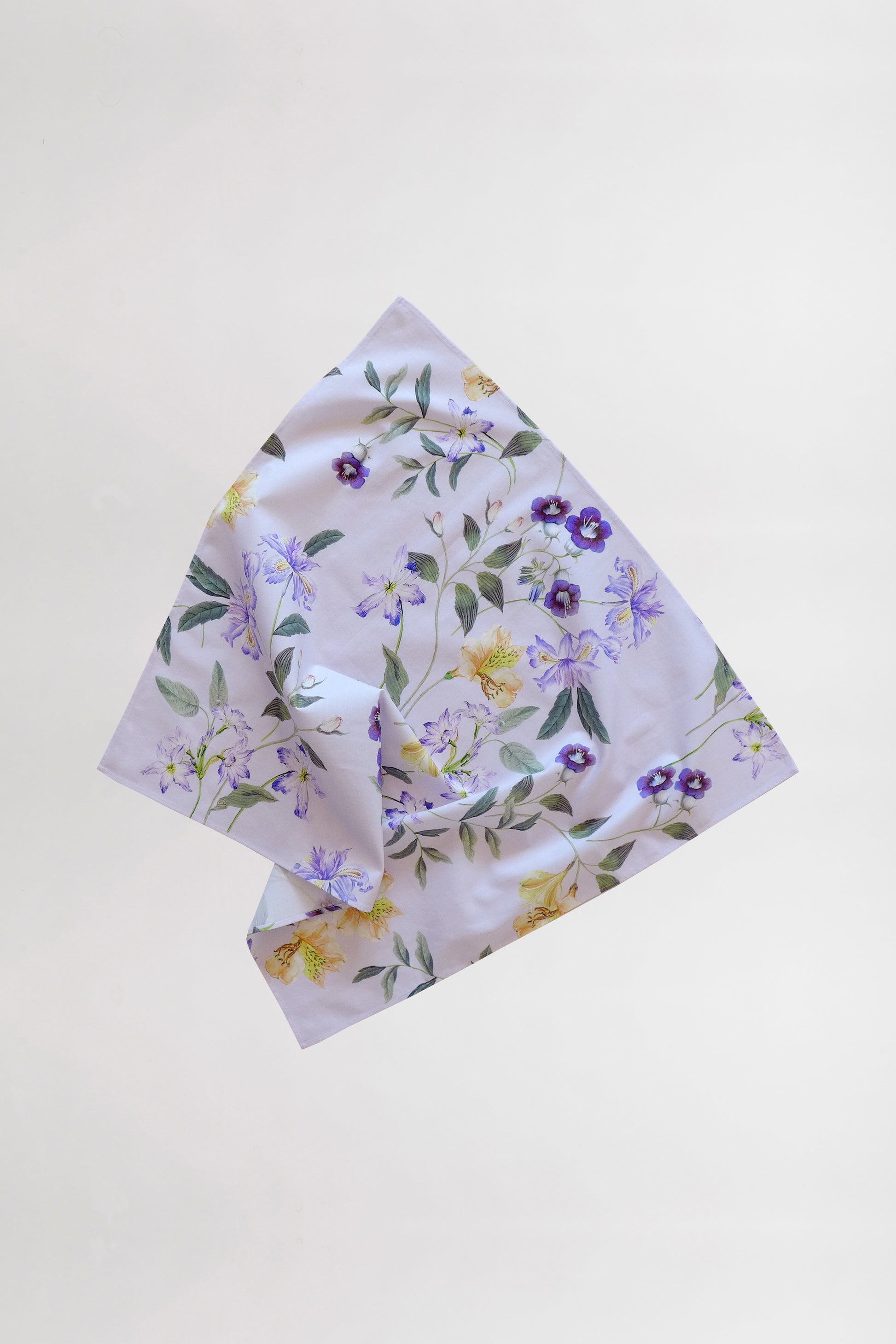 A light-colored fabric handkerchief, crafted from leftover fabric, showcases a delicate floral pattern against a plain white background. The flowers in shades of purple and yellow are complemented by green leaves interspersed among them. The fabric is slightly folded in the center.