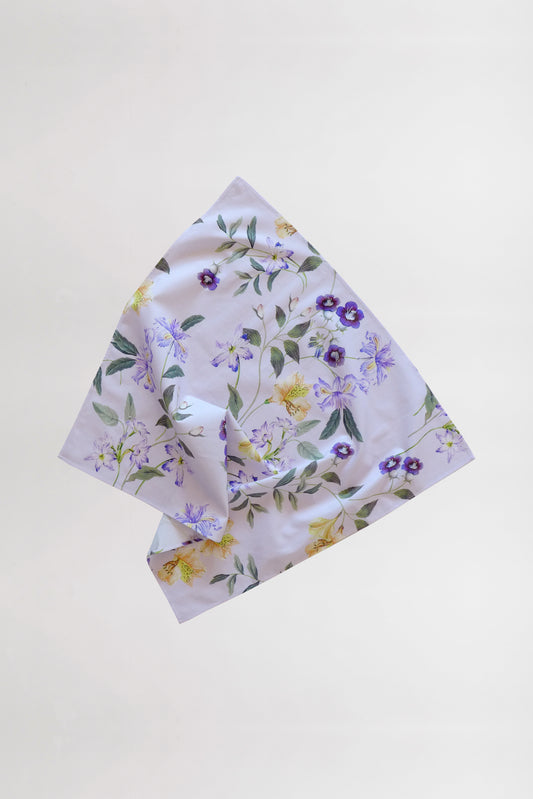 A light-colored fabric handkerchief, crafted from leftover fabric, showcases a delicate floral pattern against a plain white background. The flowers in shades of purple and yellow are complemented by green leaves interspersed among them. The fabric is slightly folded in the center.