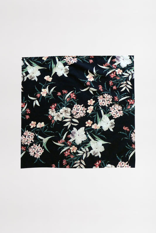 A square piece of upcycled fabric with a black background featuring an intricate floral pattern. The design includes white, pink, and light orange flowers with green leaves, creating a vibrant and elegant appearance. The foulard is laid flat against a white surface.