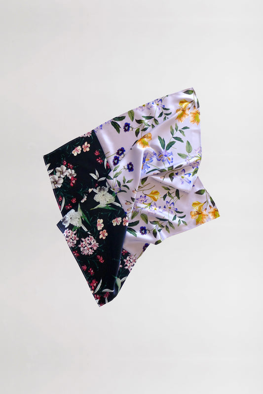 A square silk scarf with a vibrant mix and match floral prints pattern. The scarf is split into two contrasting sections: one side has a black background with white, pink, and green flowers, while the other side showcases a white background with blue, yellow, and green flowers.