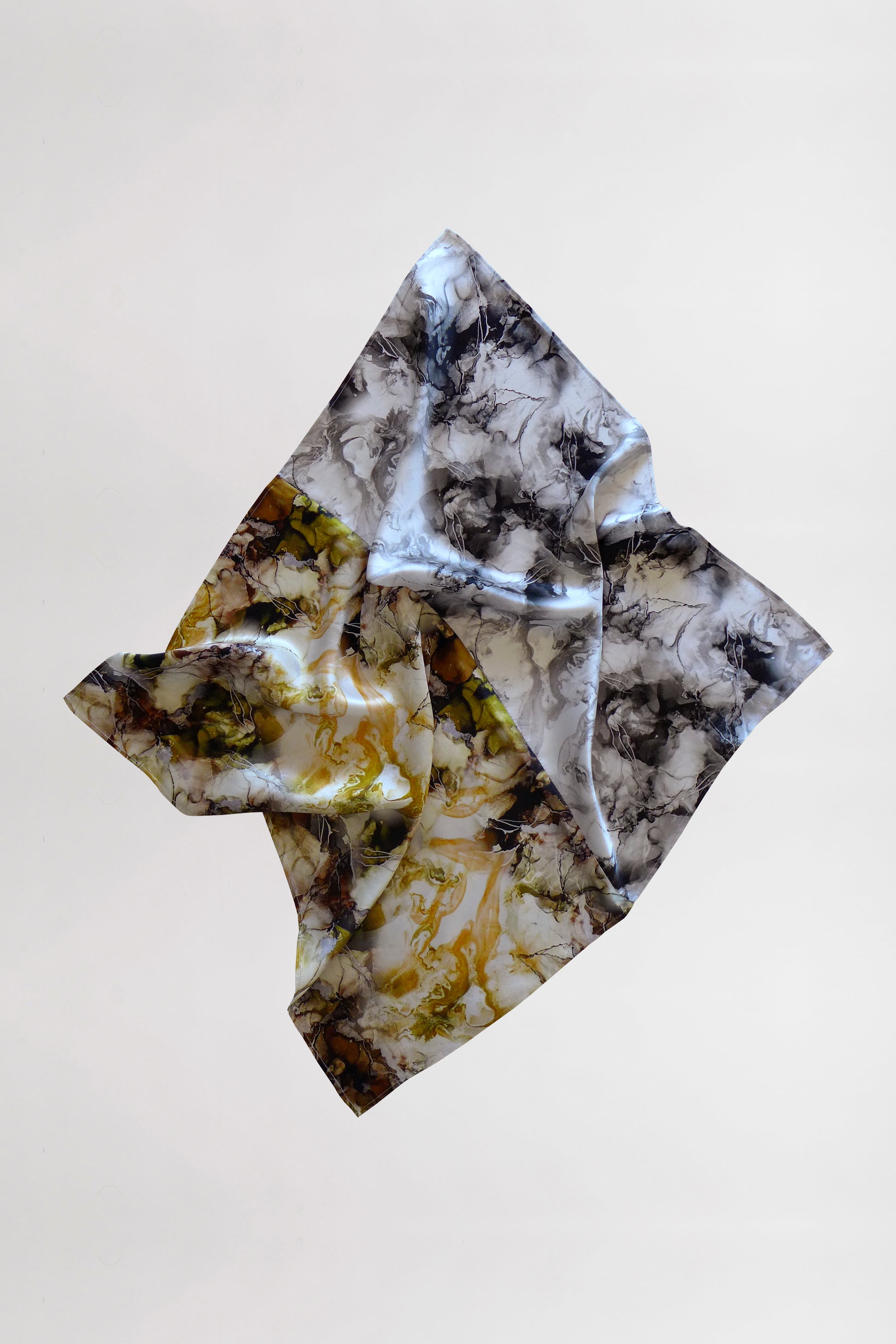 A silk scarf with a marble print in shades of gray, black, white, and yellow is captured as it floats against a plain white background. The fabric folds gracefully, creating intricate shapes and shadows.