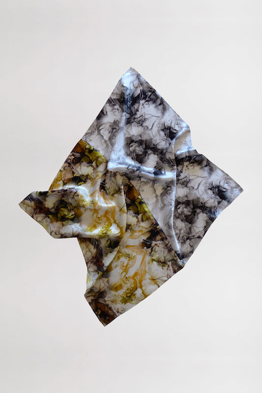 A silk scarf with a marble print in shades of gray, black, white, and yellow is captured as it floats against a plain white background. The fabric folds gracefully, creating intricate shapes and shadows.