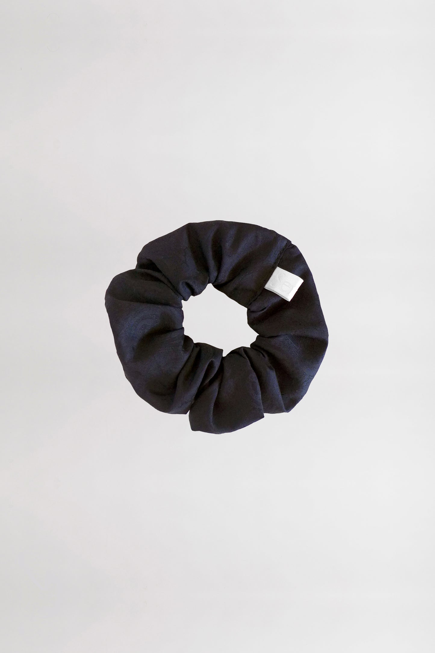 A black fabric scrunchie with a white tag is placed on a plain, light gray background. This circular accessory, made from leftover jacquard cotton fabric, has a gathered and slightly wrinkled appearance.