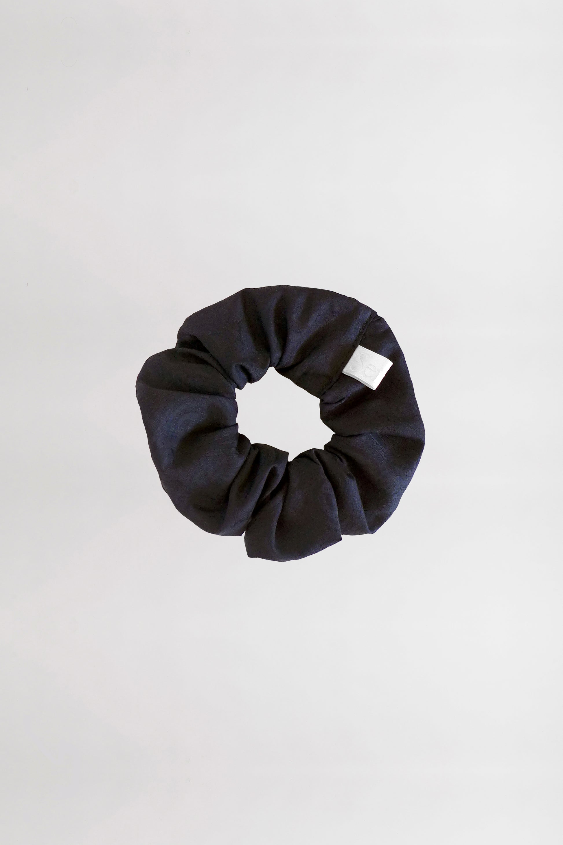 A black fabric scrunchie with a white tag is placed on a plain, light gray background. This circular accessory, made from leftover jacquard cotton fabric, has a gathered and slightly wrinkled appearance.