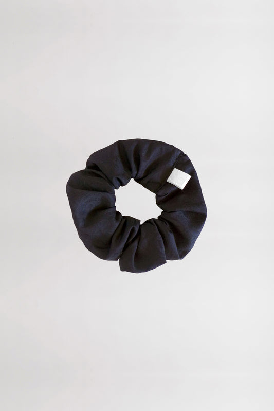 A black fabric scrunchie with a white tag is placed on a plain, light gray background. This circular accessory, made from leftover jacquard cotton fabric, has a gathered and slightly wrinkled appearance.