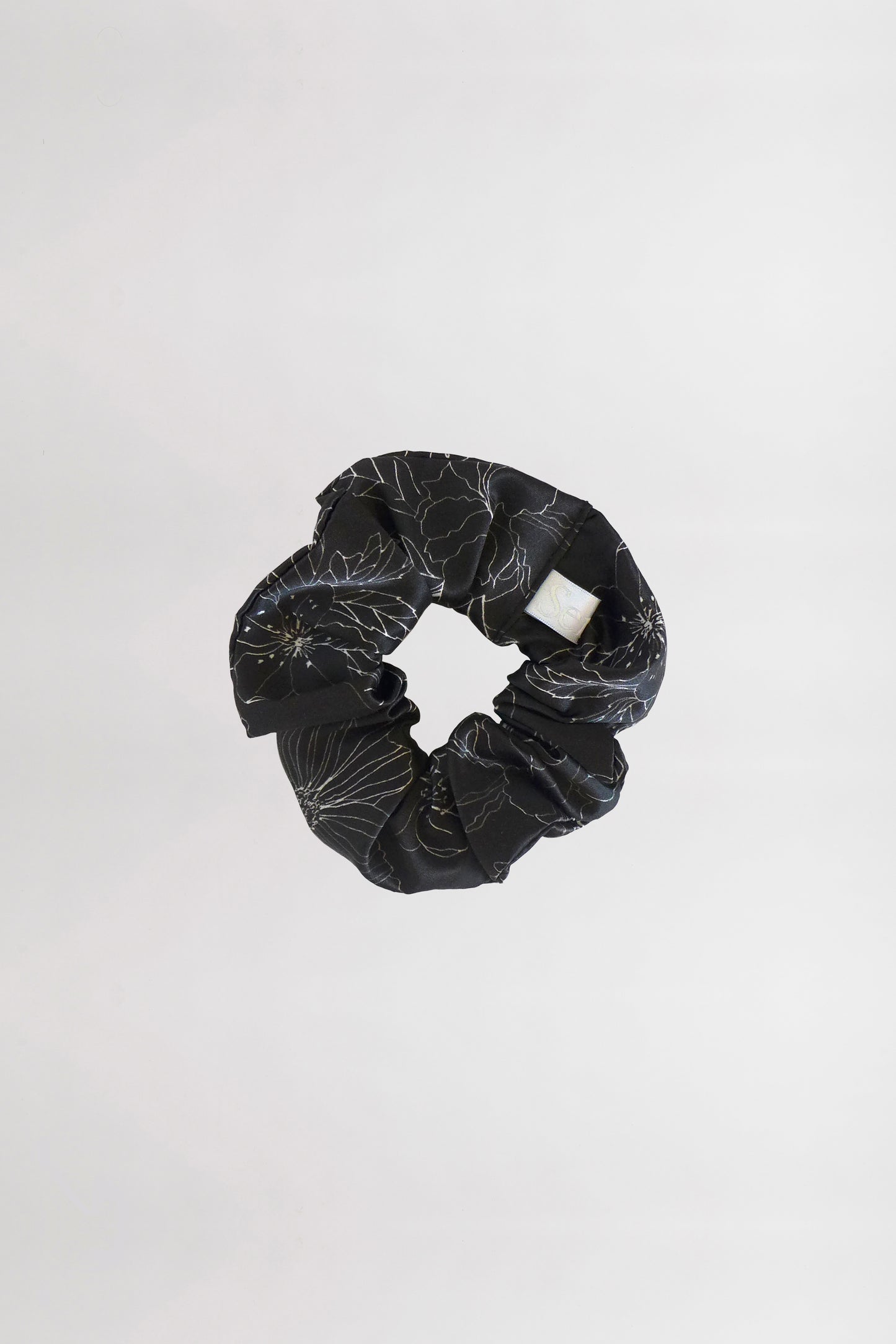 A black fabric scrunchie with a subtle linear flower print and a white brand tag is centered against a plain white background. Made from leftover fabric, the scrunchie is arranged in a circular shape, showcasing its gathered texture.