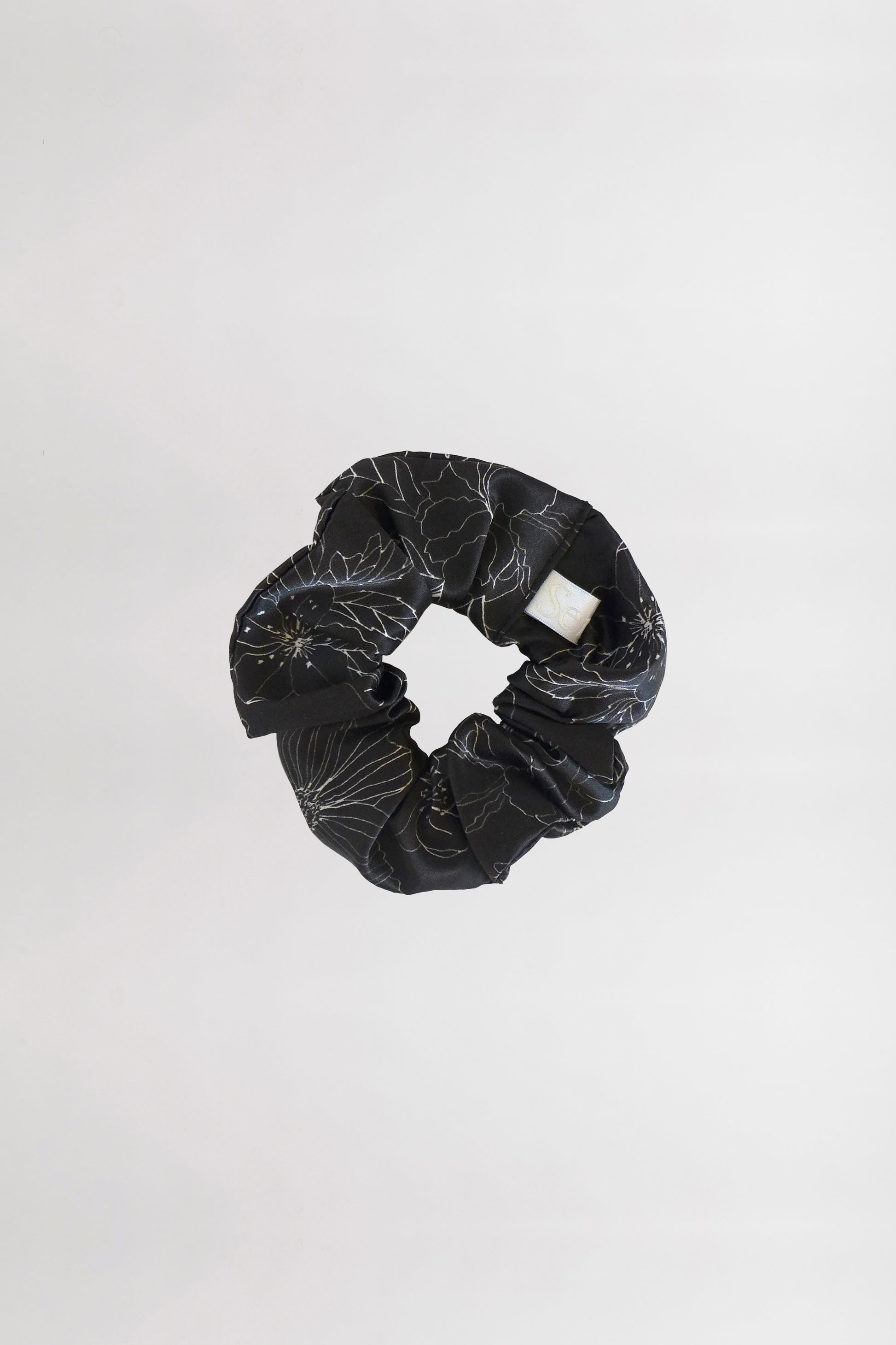 A black fabric scrunchie with a subtle linear flower print and a white brand tag is centered against a plain white background. Made from leftover fabric, the scrunchie is arranged in a circular shape, showcasing its gathered texture.