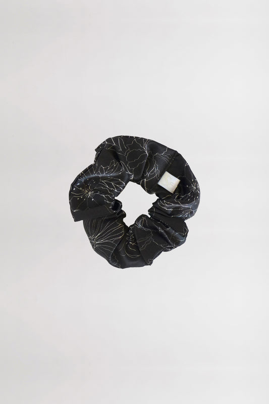 A black fabric scrunchie with a subtle linear flower print and a white brand tag is centered against a plain white background. Made from leftover fabric, the scrunchie is arranged in a circular shape, showcasing its gathered texture.