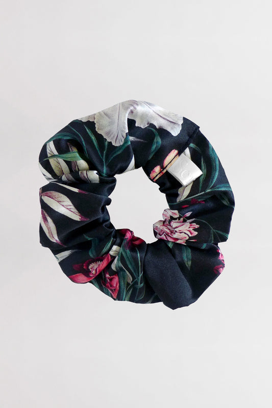 A silk satin scrunchie with a floral pattern featuring white, pink, and green colors, displayed against a light gray background. The scrunchie is arranged in the center, showcasing its fabric texture and vibrant tropical flower print on a dark background.