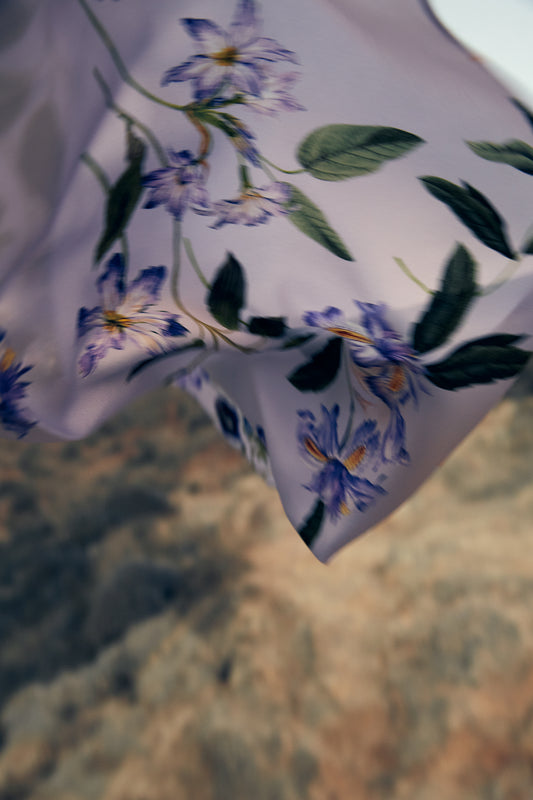 A light purple scarf with a floral pattern featuring purple flowers and green leaves, made from leftover fabric, gently blows in the wind against a blurred, rocky background.