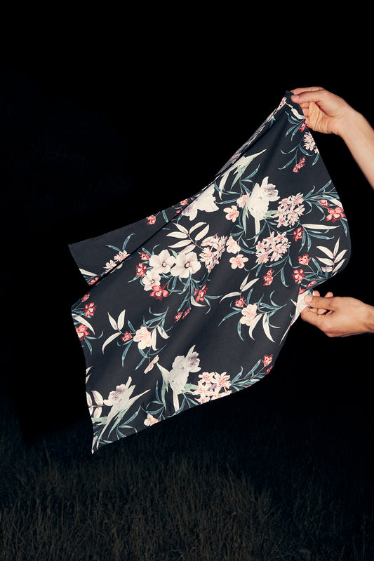 Two hands hold up a dark cotton foulard with a vibrant floral pattern featuring white, pink, and red tropical flowers along with green leaves. The background is completely black, highlighting the foulard-inspired design.