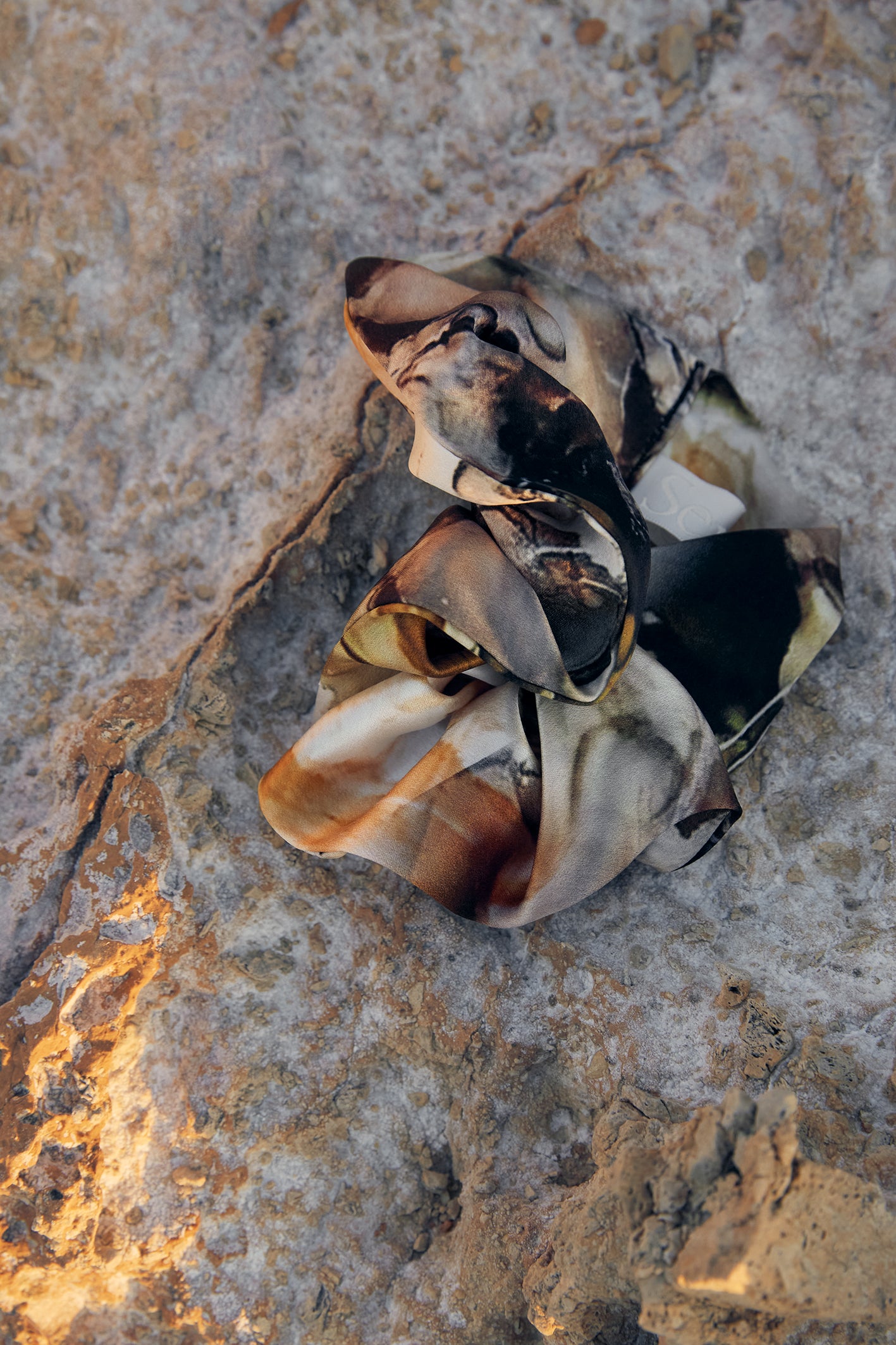 A crumpled, multi-colored silk satin fabric scrunchie with earthy tones lies on a textured, rocky surface. Warm light illuminates part of the fabric and the surrounding rocky ground, enhancing the rugged texture.