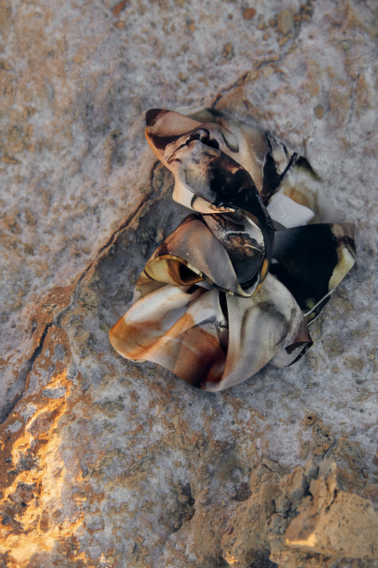 A crumpled, multi-colored silk satin fabric scrunchie with earthy tones lies on a textured, rocky surface. Warm light illuminates part of the fabric and the surrounding rocky ground, enhancing the rugged texture.