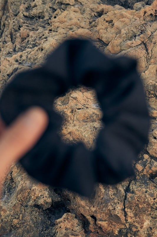 A black hair scrunchie made from leftover fabric is held up against a textured, rocky background. The scrunchie appears out of focus while the rocks are in clear detail, showcasing their rough and uneven surface.