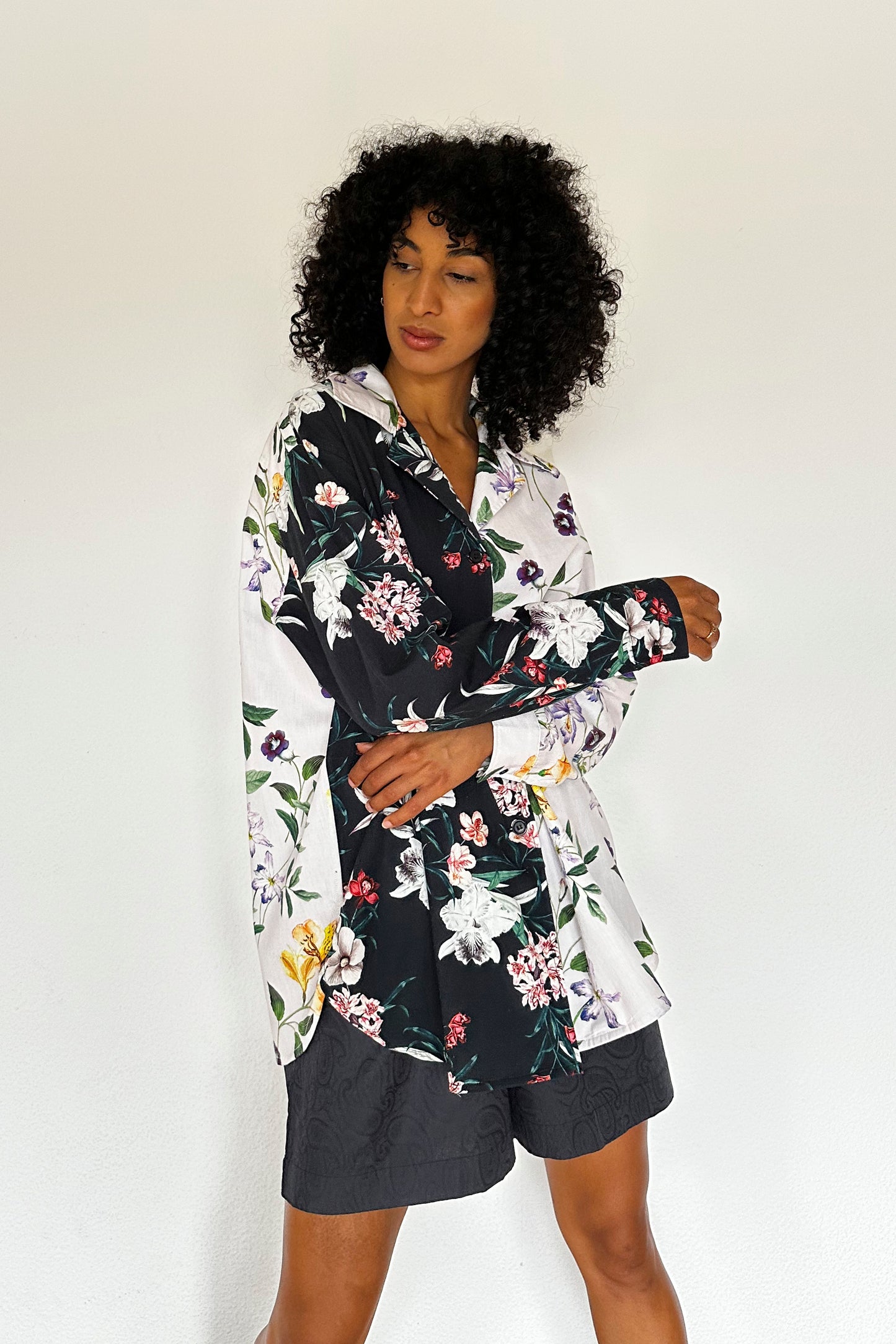 A person with curly hair stands against a plain white background, wearing a loungewear shirt with a mixed floral pattern in black and lilac and textured jacquard shorts. Their arms are relaxed, and they are looking slightly downward with a calm expression.