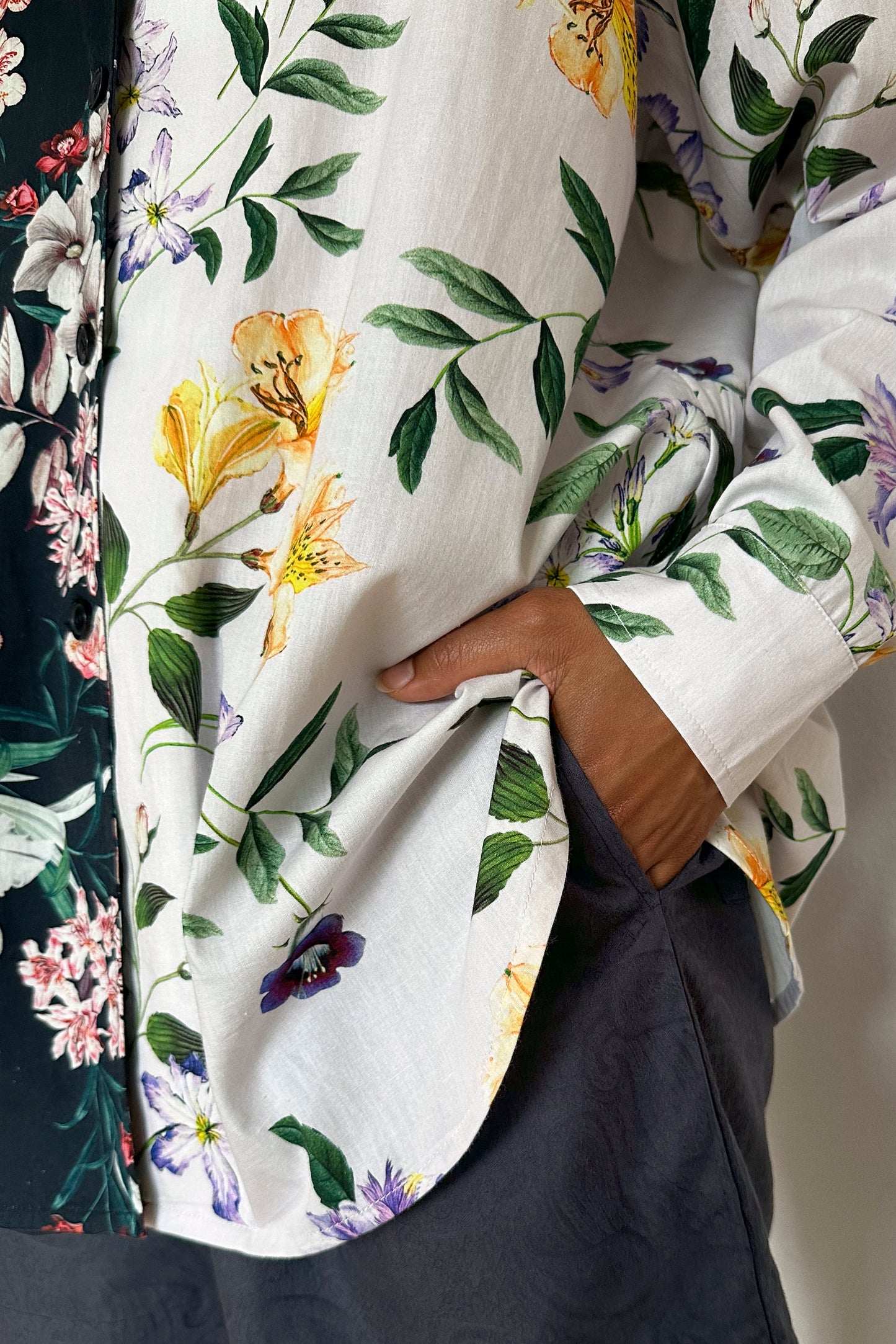 A person stands with one hand in their pocket, wearing dark shorts and a mix and match printed floral pattern loungewear shirt that features detailed yellow and purple flowers with green leaves. The image captures a casual, stylish look.