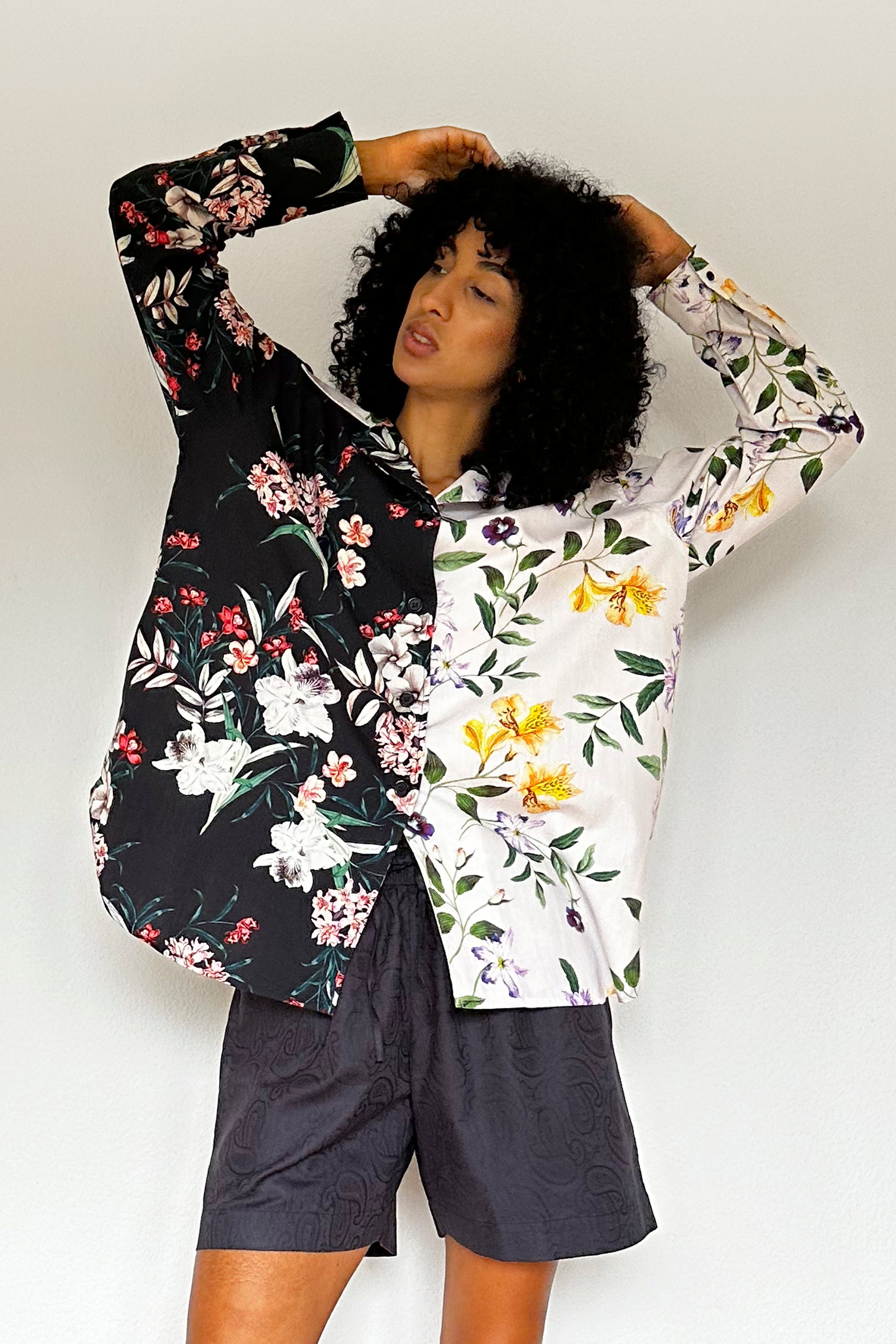 A person with curly hair is posing against a plain white background. They are wearing a long-sleeved shirt featuring a mix and match design; one half is black with colorful flower prints, while the other half is lilac with different floral patterns. They also sport dark shorts made of a textured fabric with cotton.