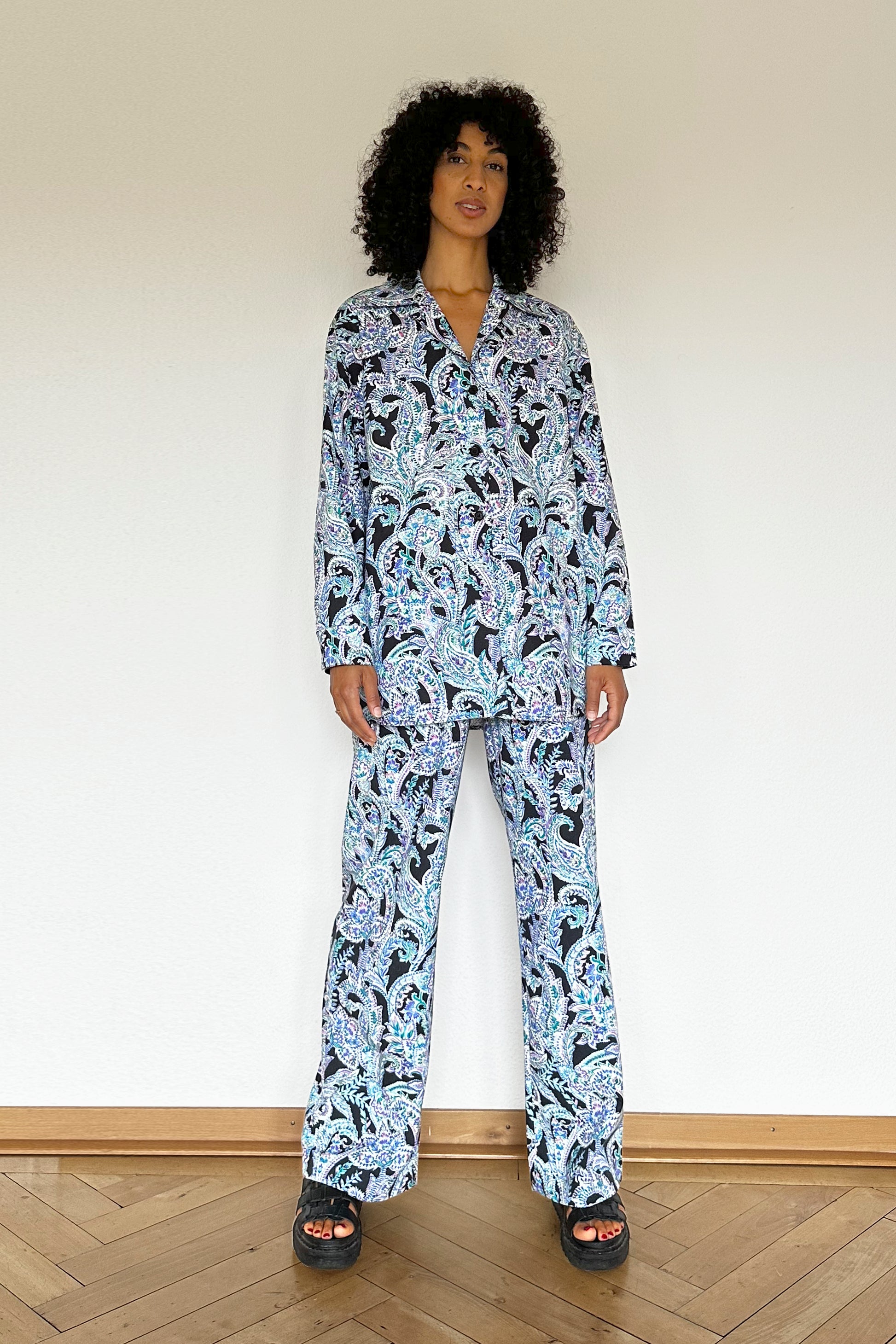 A person with curly hair is standing against a blank wall, wearing a coordinated loungewear set comprised of a paisley-patterned long-sleeve shirt and matching pants. The model is also wearing black platform sandals.