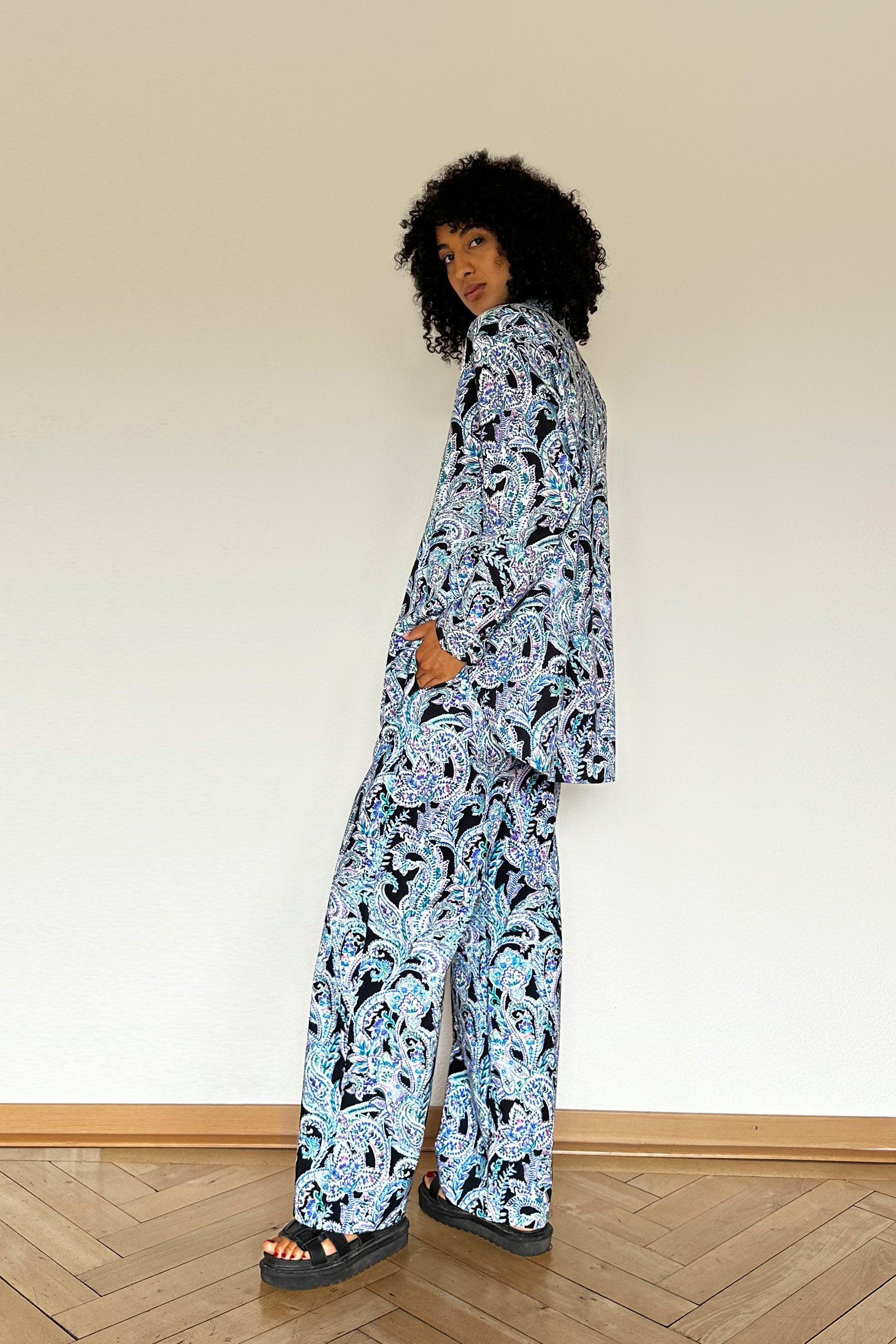 A person with curly hair stands against a plain white wall, wearing a matching set of a paisley-patterned shirt and wide-legged pants in shades of blue, black, and white. Black platform sandals complete the look. The person looks towards the camera over their shoulder.