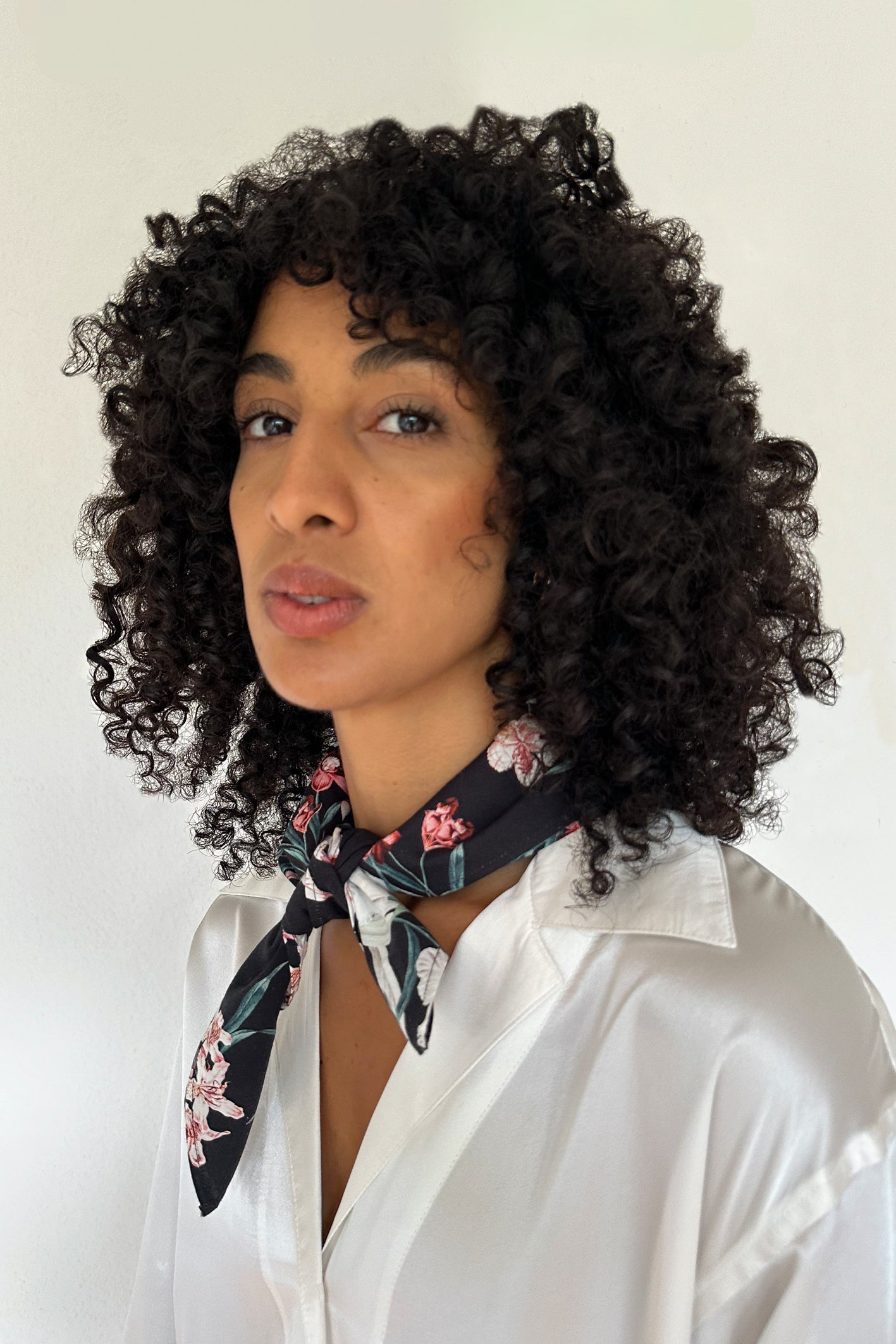 A person with curly black hair wears a white shirt and a bandana made from leftover fabric knotted around their neck. They are looking directly at the camera with a neutral expression. The background is plain and white.