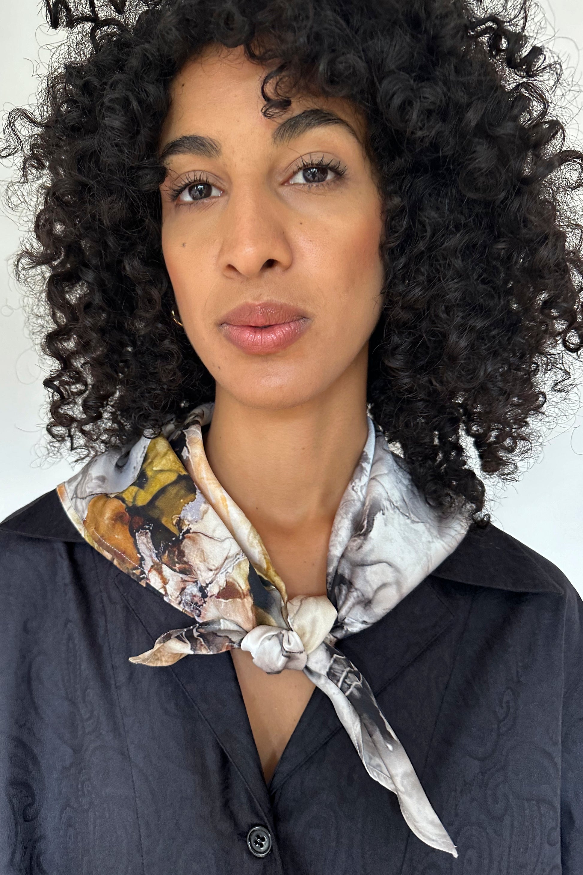 A person with curly hair is wearing a black buttoned shirt and a patterned silk scarf knotted at the neck, effortlessly showcasing a mix-and-match style. The person is looking directly at the camera with a neutral expression, set against a plain white background.