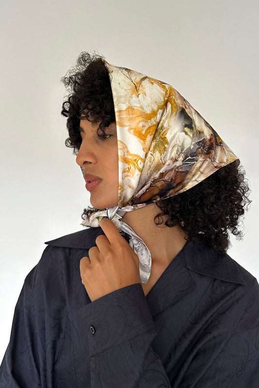 A person with curly hair is wearing a marble-patterned silk scarf, upcycled from leftover fabric, wrapped around their head and tied under the chin. The individual dons a dark, textured shirt and gazes to the side against a light, plain background.