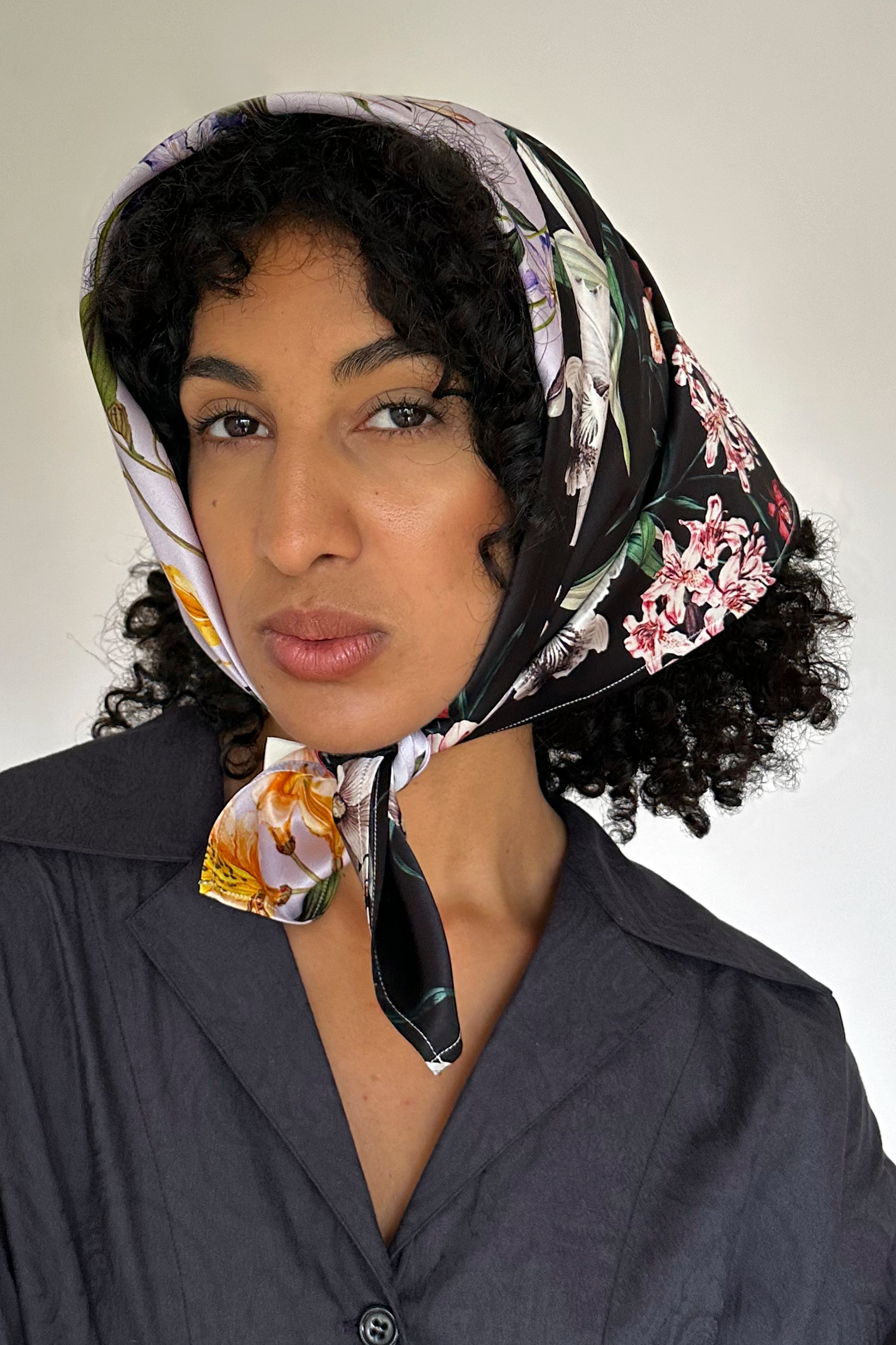 A person with curly hair wearing a colorful floral print scarf tied under the chin and a dark button-up shirt gazes into the camera against a plain background.