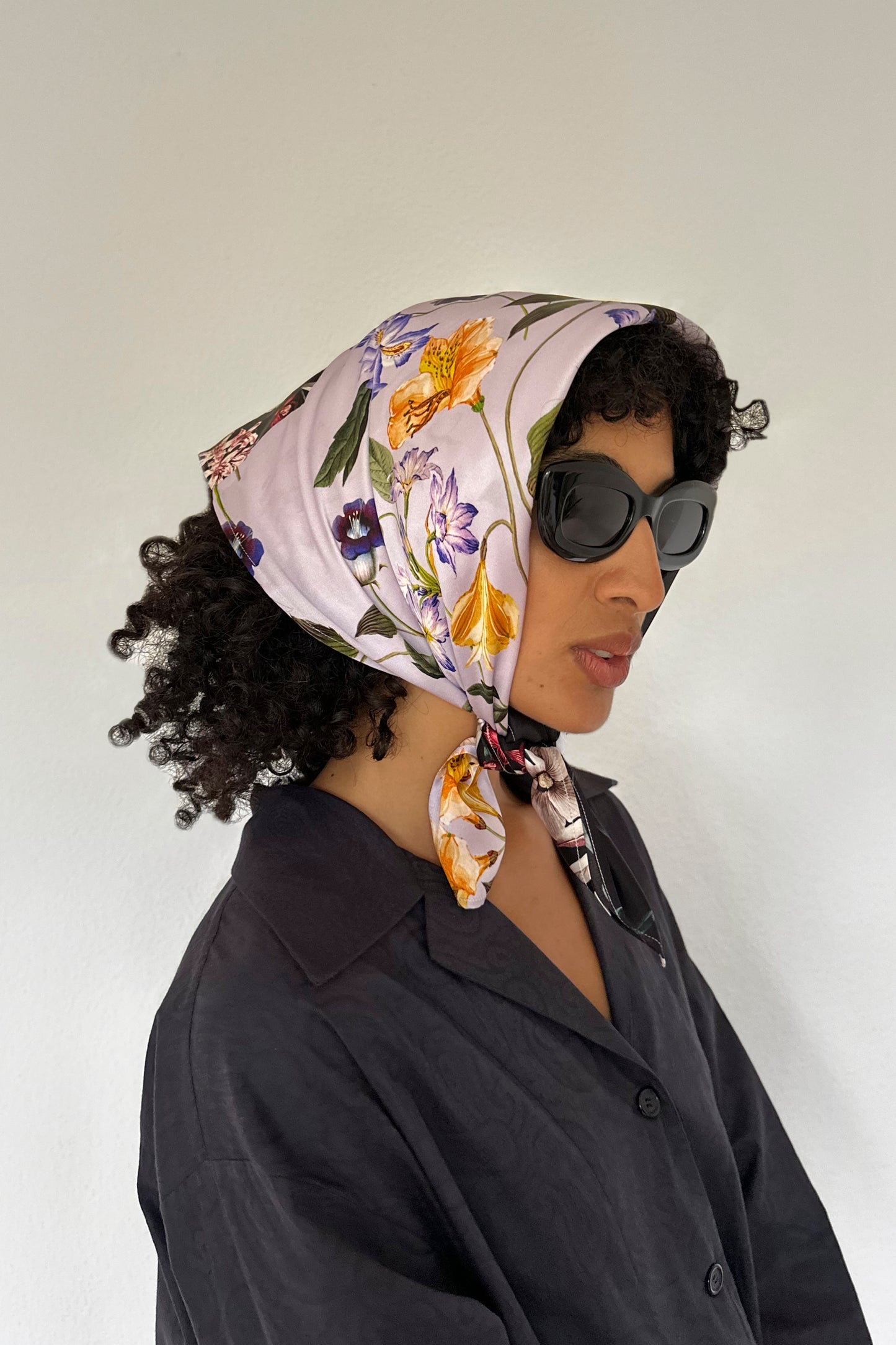 A person with curly hair, wearing dark sunglasses and a silk scarf tied over their head, is seen in profile against a plain background. The floral print of the scarf adds a touch of elegance to their dark button-up shirt.