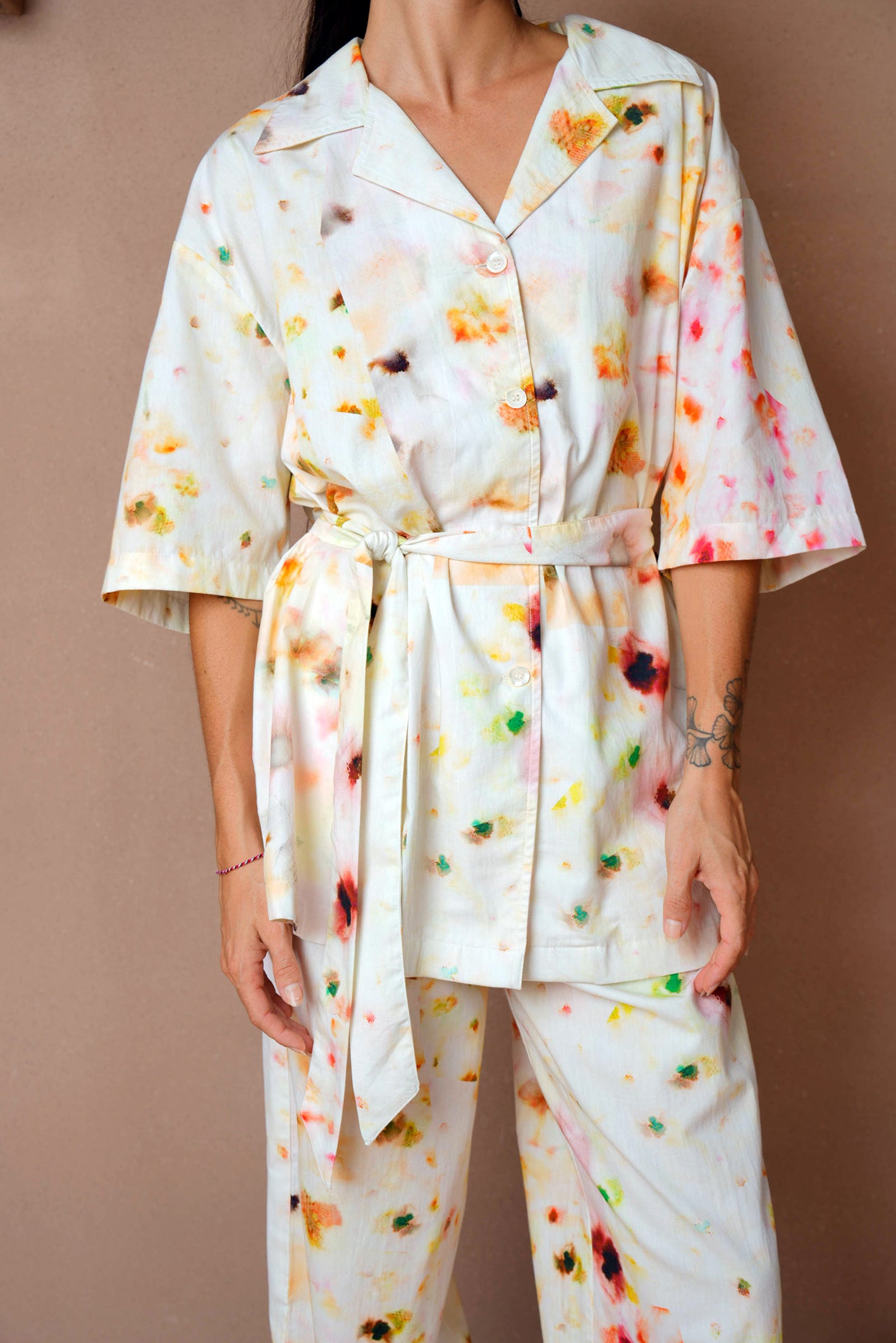 A cropped front view of a woman wearing a relaxed-fit loungewear set with an abstract floral print. The short-sleeve button-up blouse features a waist belt, and the matching wide-leg pants complete the look. She stands against a neutral-toned background.