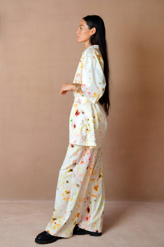 A side view of the same woman wearing the floral loungewear set, showcasing the flowy silhouette of the pajama outfit. Her long dark hair cascades down her back as she stands against the same neutral background, wearing black loafers.