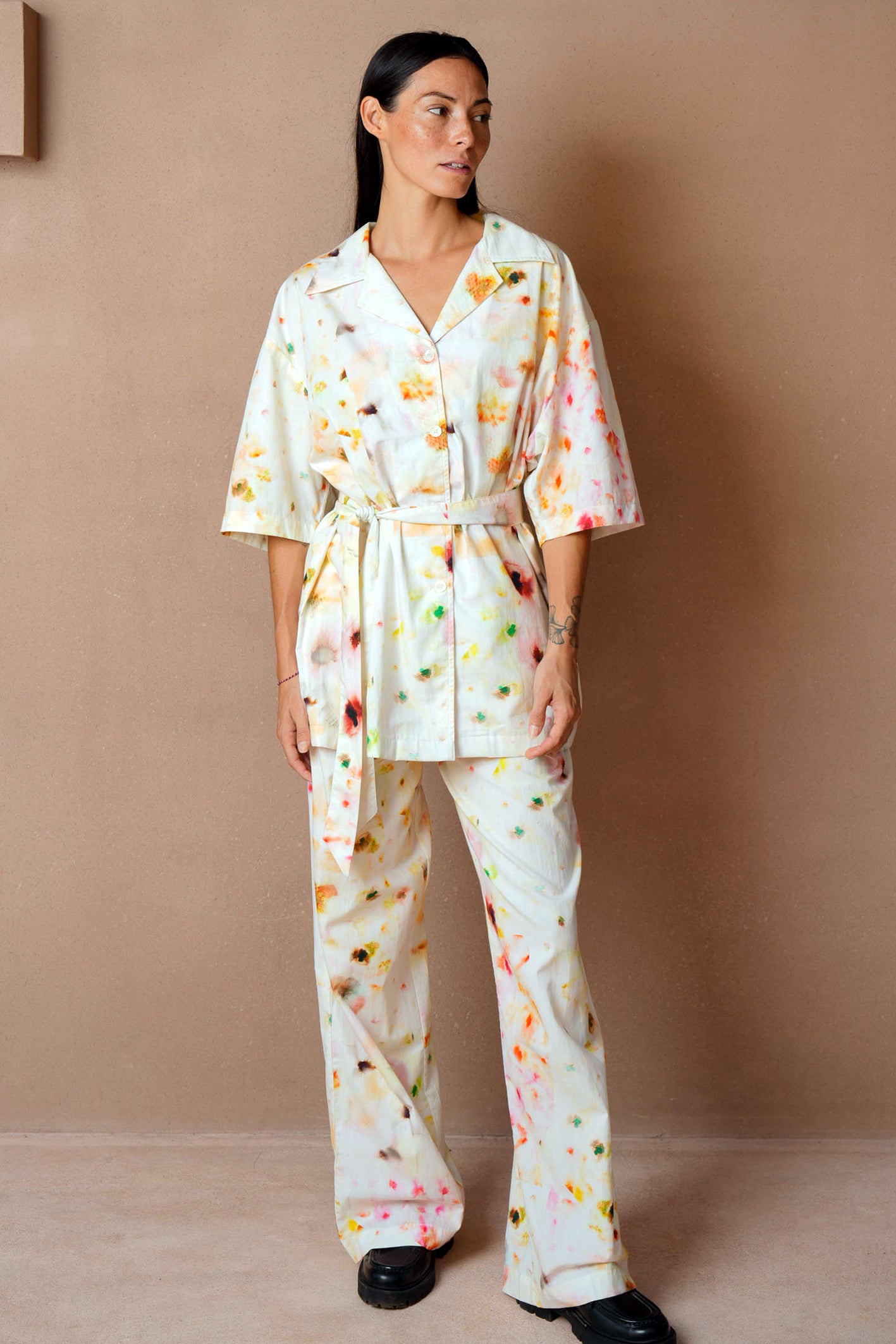 A woman wearing a relaxed-fit loungewear set with an abstract floral print in soft pastel tones. The outfit features a short-sleeve button-up top with a waist belt and wide-leg pants, styled with black loafers. She stands against a neutral-toned background, looking off to the side.
