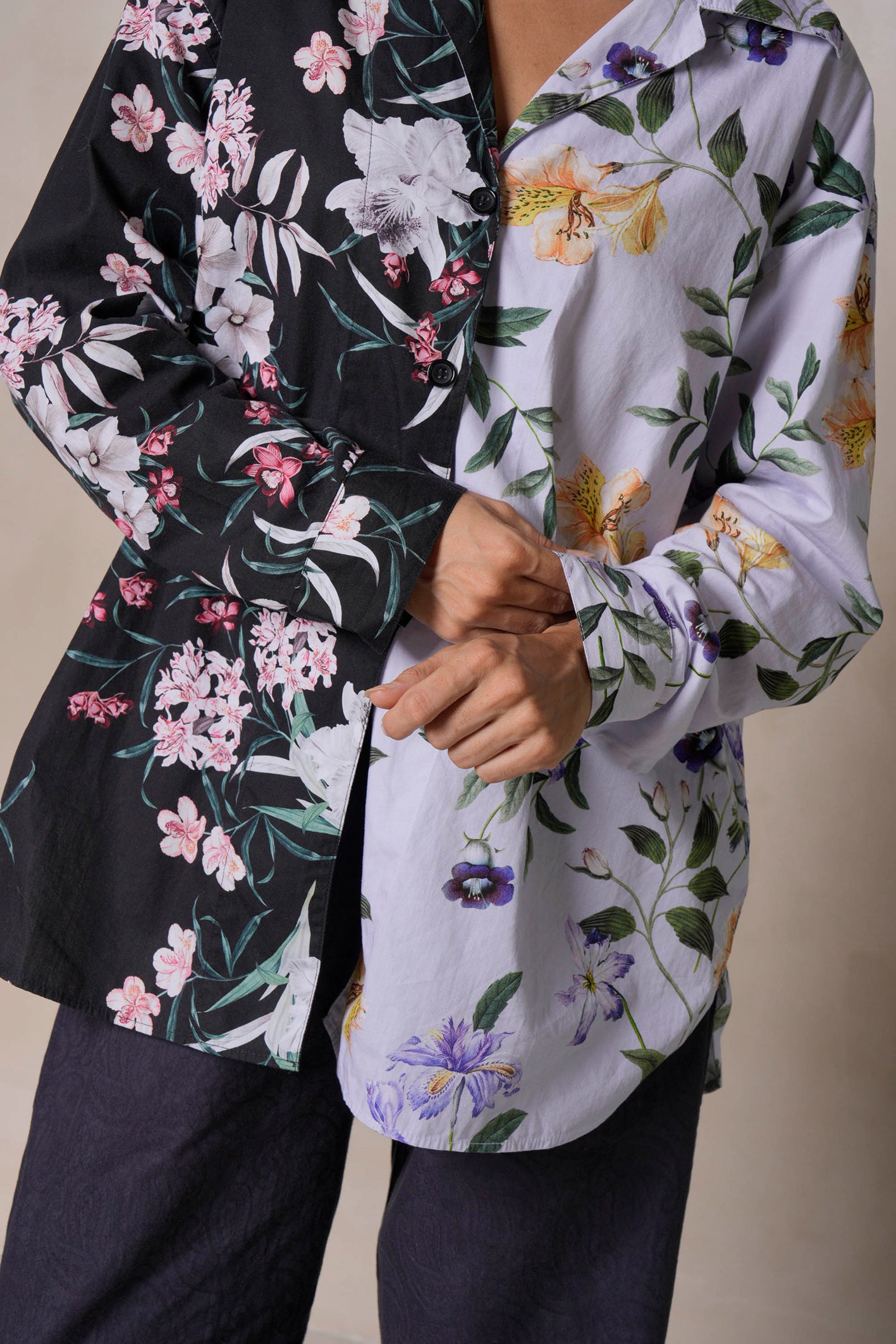 A close-up of a woman's hands adjusting the cuff of her blouse, highlighting the intricate tropical flower print in black and lilac. The organic cotton fabric contrasts beautifully with the dark jacquard paisley pants, showcasing luxurious loungewear details.