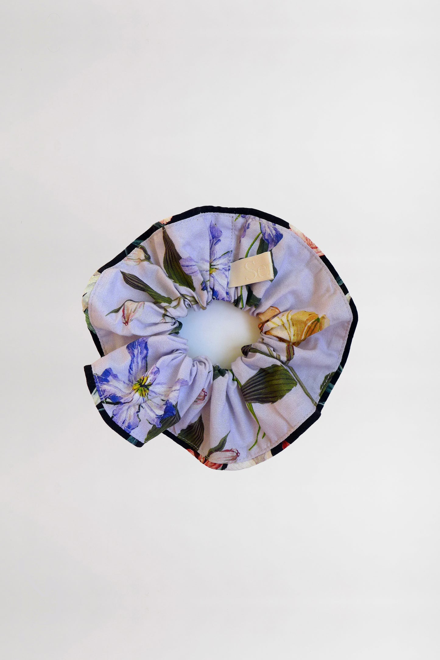 A floral-patterned scrunchie with contrasting black piping made from leftover fabric, featuring delicate blue, yellow, and pink flowers on a light background. The scrunchie is displayed against a plain white backdrop.