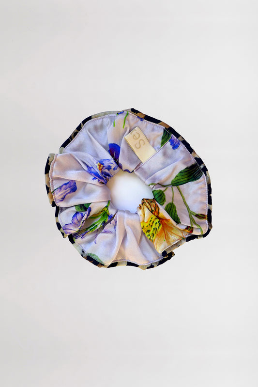 A floral-patterned scrunchie with a contrasting piping in a leopard print. The fabric features delicate illustrations of flowers and leaves in shades of yellow, green, and purple. A small beige tag is attached to this charming scrunchie. The background is plain white.