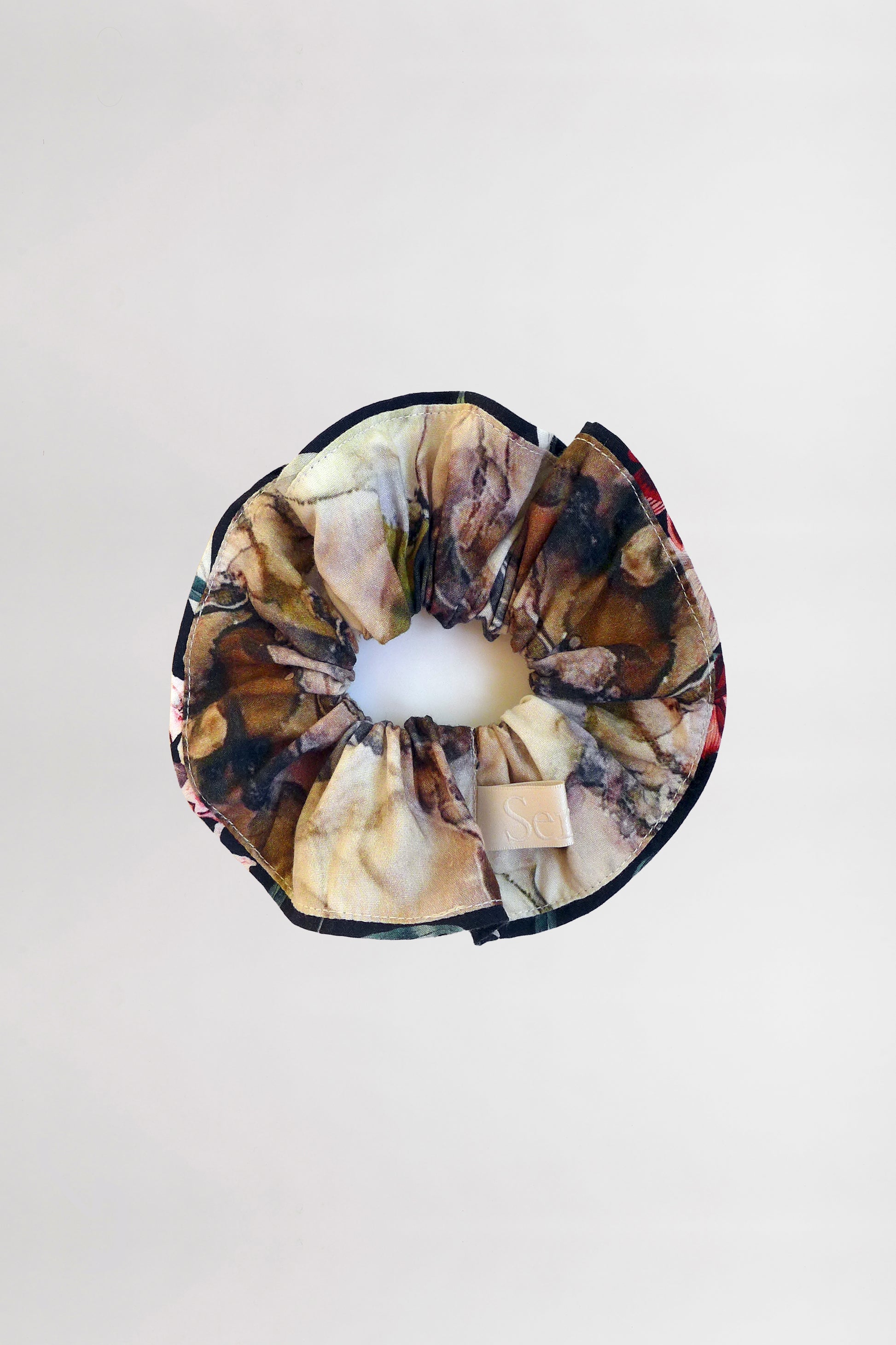 A scrunchie made from leftover fabric in a marble-like pattern of brown, black, and beige with contrasting black piping. This organic cotton scrunchie is displayed against a plain gray background, adding an eco-friendly touch to your style.