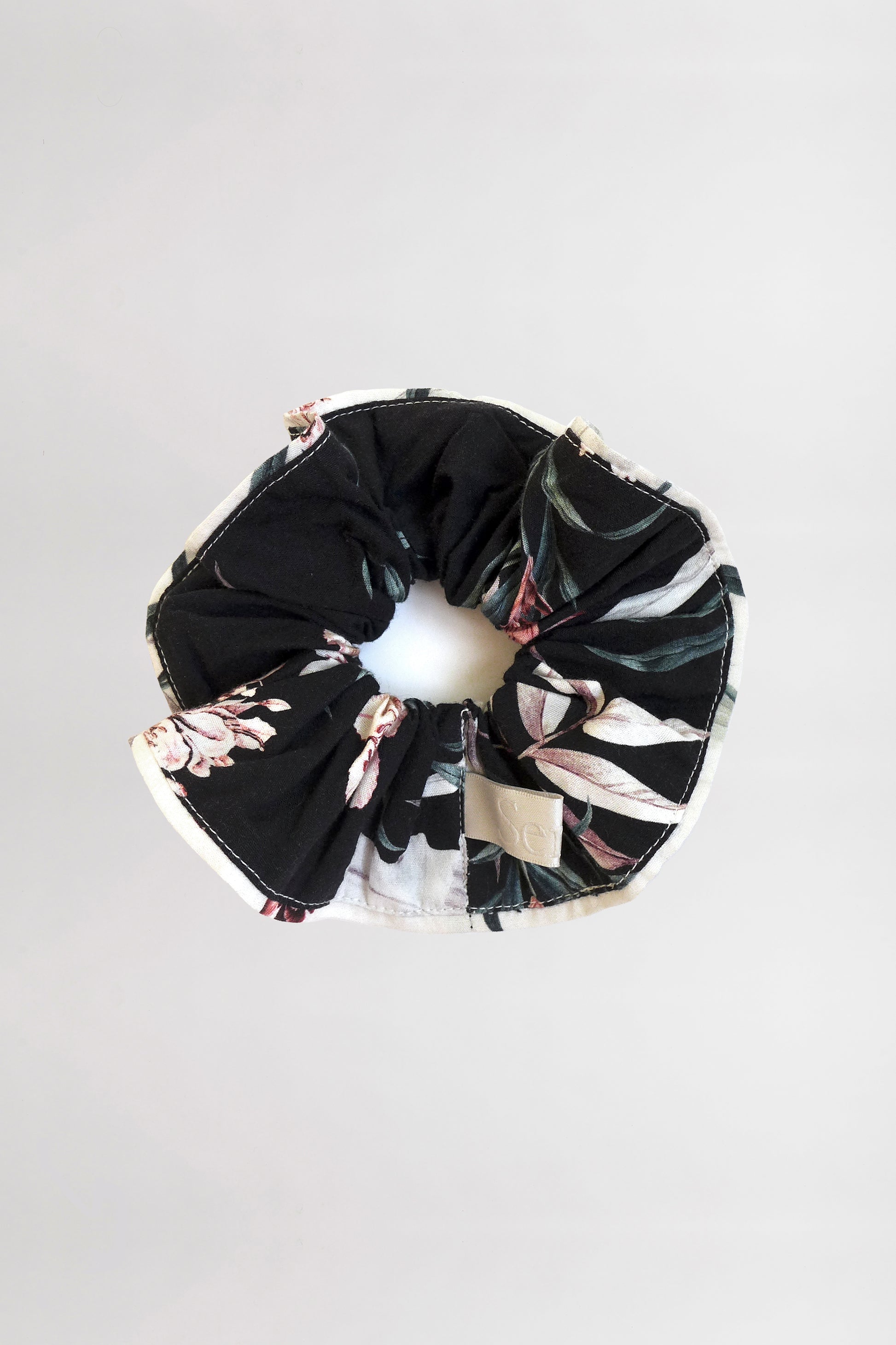 A close-up image of an Organic Cotton Scrunchie with a floral pattern on a black background with contrasting cream piping. Made from leftover fabric, the scrunchie is positioned on a plain, light-colored surface.