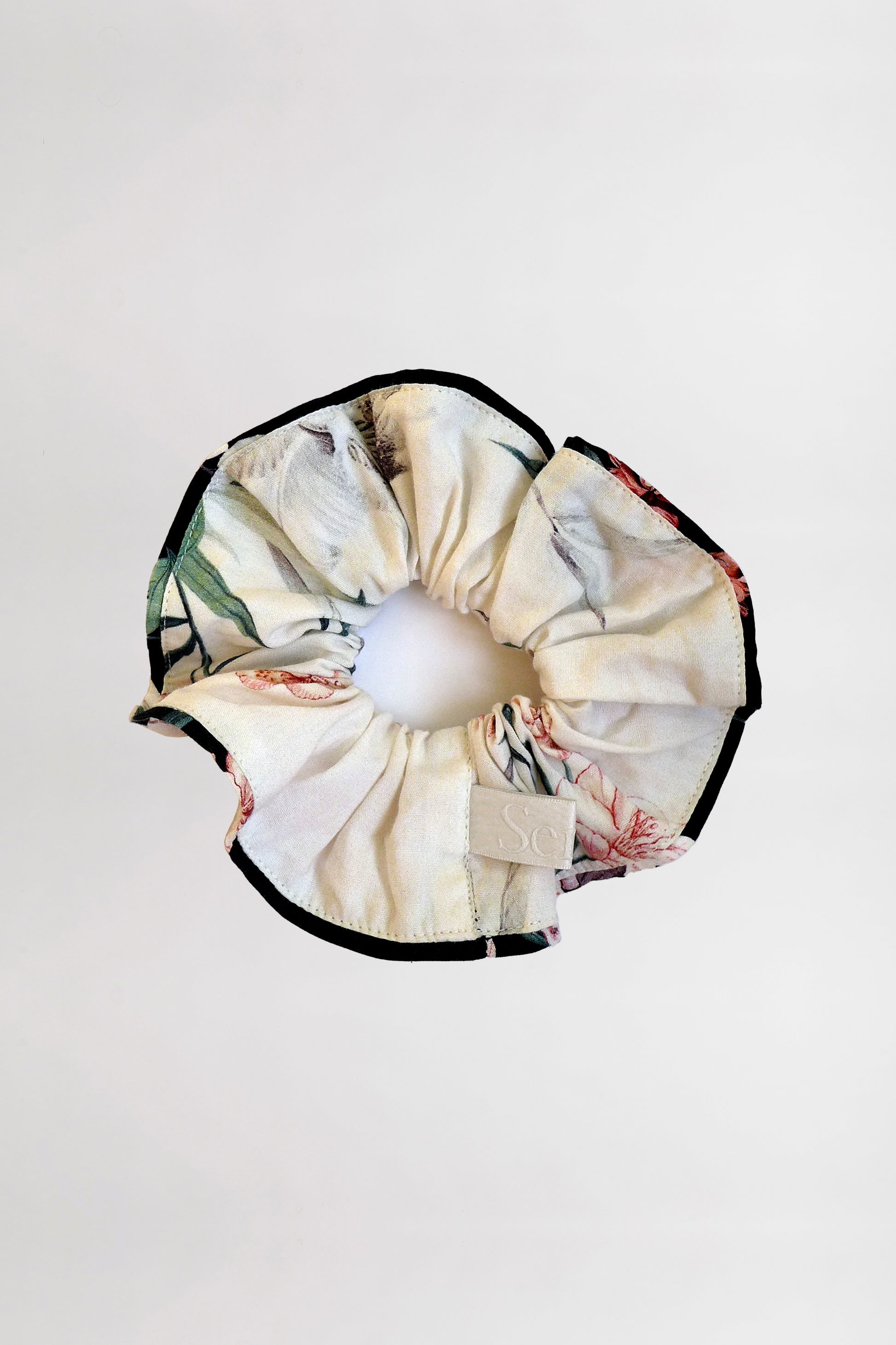 A soft scrunchie made from organic cotton with a multicolored floral print, featuring hues of beige, green, and red with contrasting black piping. The scrunchie also has a small, rectangular light beige tag stitched into it. Crafted from leftover fabric, the background is plain white.