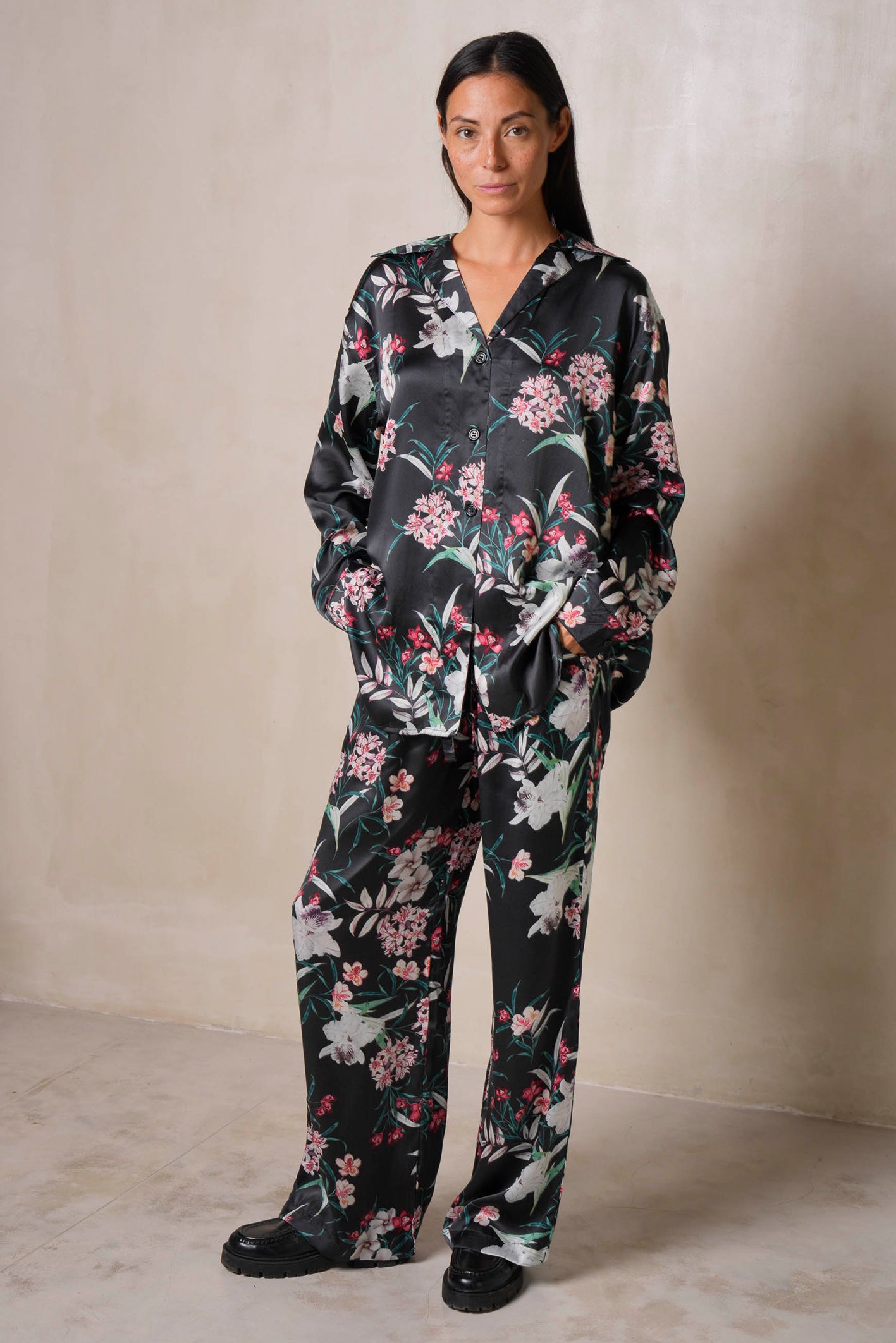 A woman stands against a neutral wall wearing a black silk loungewear set with long sleeves. The outfit features tropical flower print patterns in pink and white. They have their hands in their pockets and are wearing black shoes.