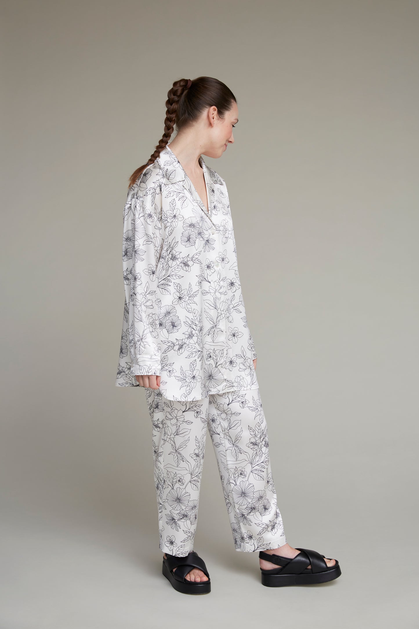 Woman in left-facing white floral-print loungewear pajama set, paired with black sandals, against neutral background, with long braid.