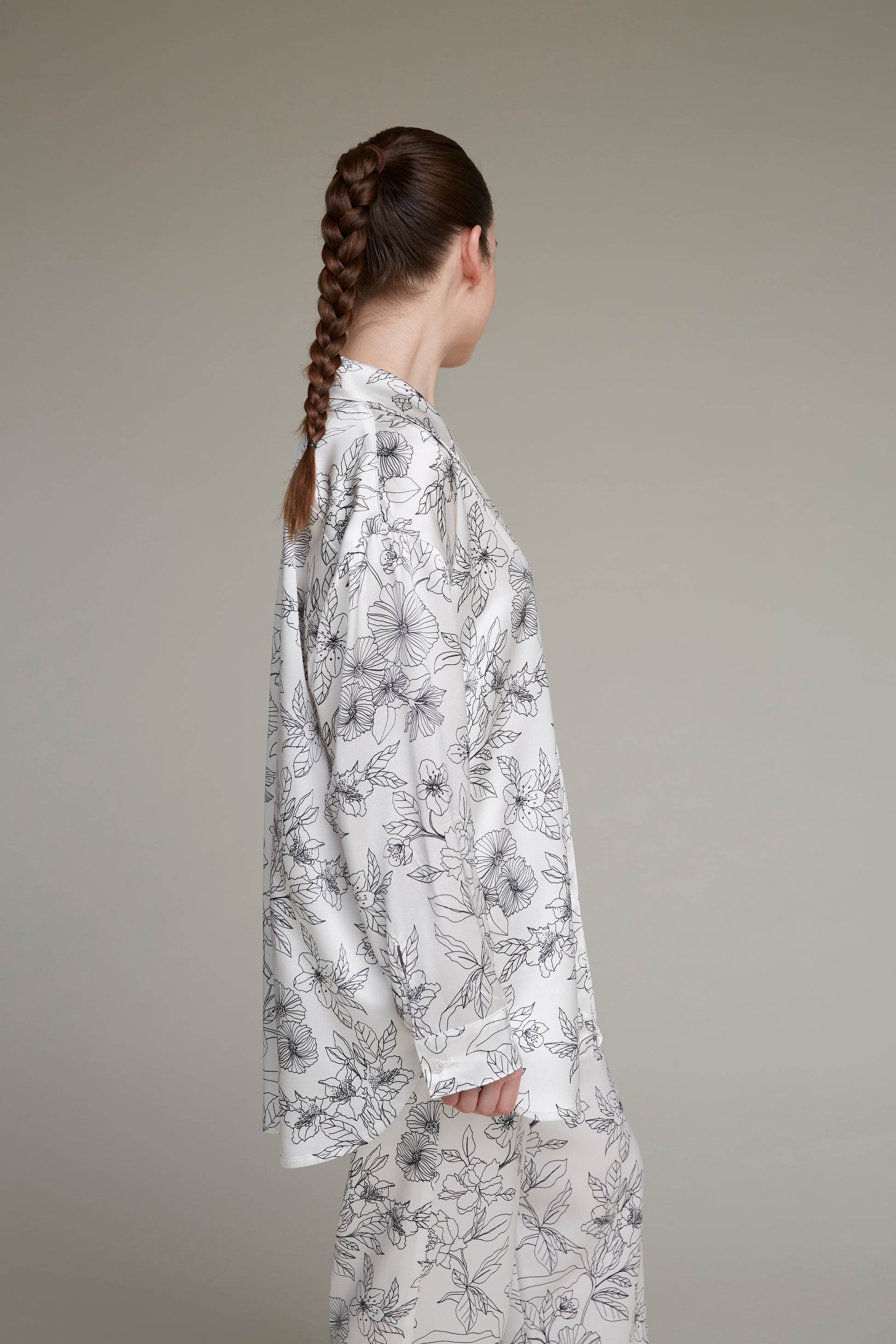A woman with a long braid wearing a white floral-print loungewear pajama set consisting of a blouse and pants, facing away against a neutral background.