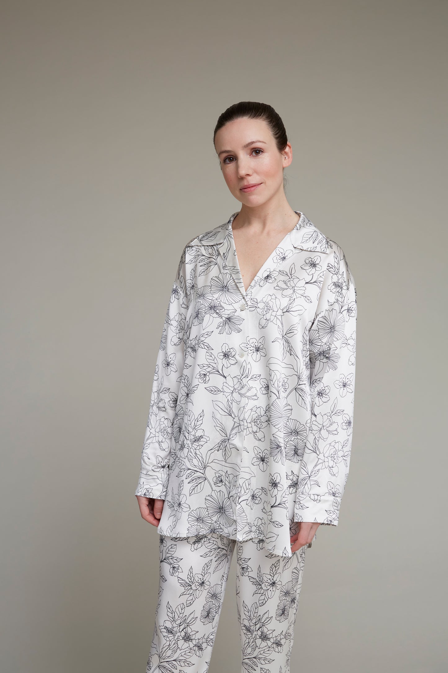 Close-up of a woman facing forward wearing a white floral-print loungewear pajama set, highlighting intricate details of the shirt and pants against a neutral background.