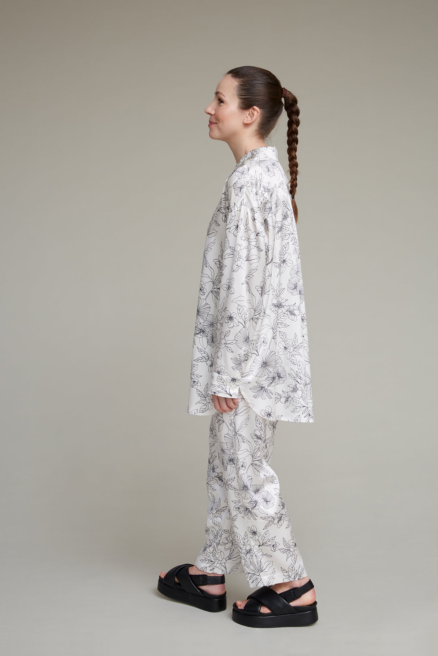 Woman in right-facing white floral-print loungewear pajama set, paired with black sandals, against neutral background, with long braid.