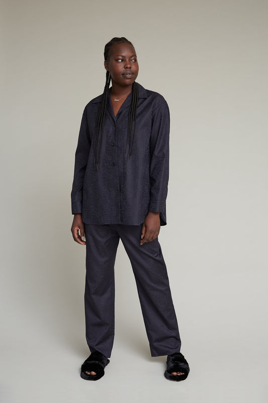 A woman in a black textured jacquard loungewear set, featuring an oversized blouse and matching pants, stands against a neutral backdrop, looking to the side.