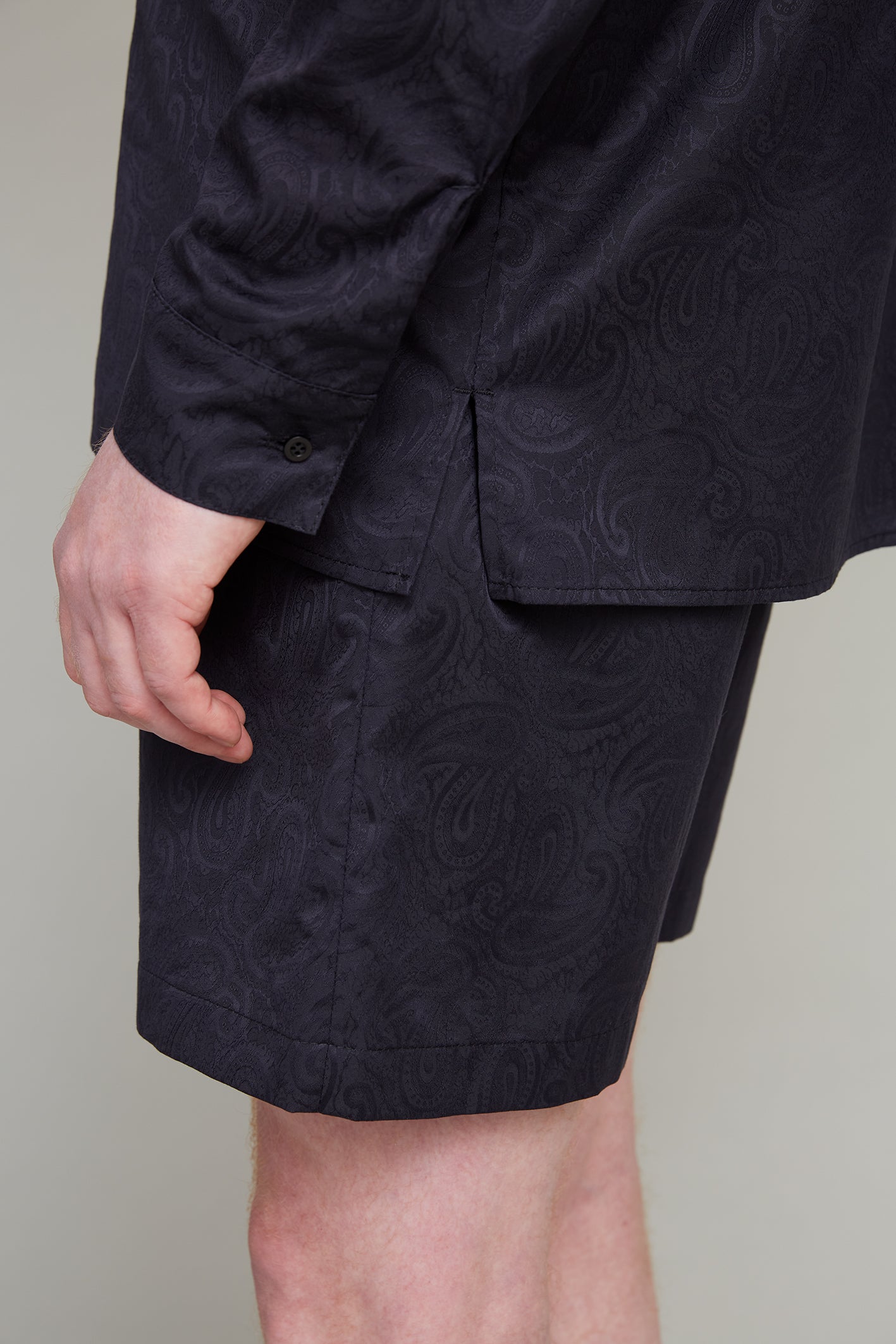 A close-up of a person wearing matching black loungewear shorts and a shirt, both made from Jacquard Paisley fabric. The person's arm rests beside their body, showcasing the intricate paisley design on both pieces. The background is plain and neutral.