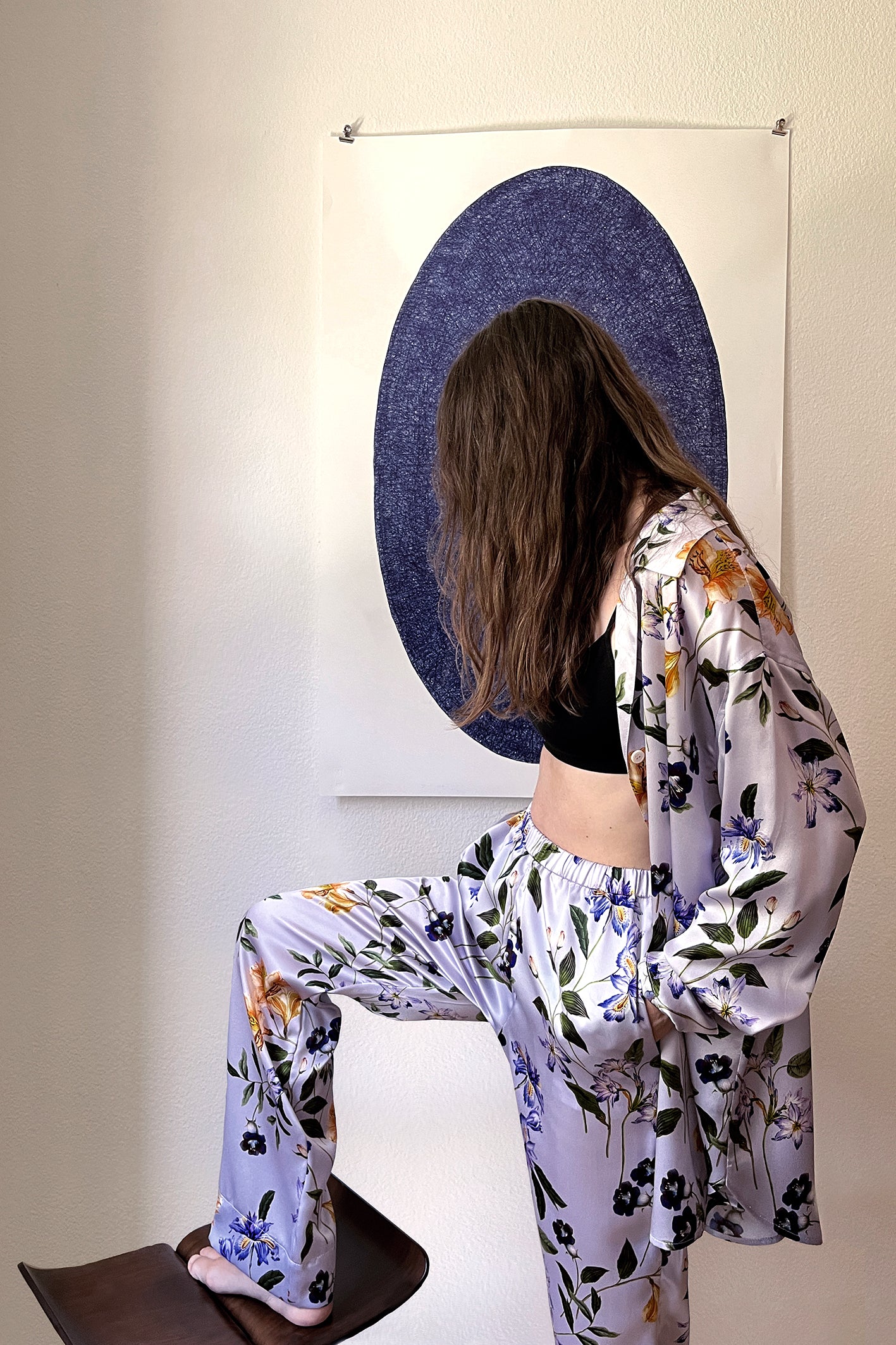 A person with long hair wearing a floral-patterned silk satin loungewear set, consisting of a shirt and matching pants, with a black top underneath. They have one foot on a chair and are looking down. Behind them is a white wall with an artwork of a dark blue oval.